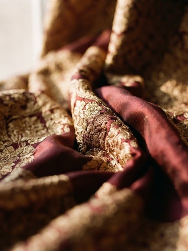 Close-up of elegant draped silk fabrics in deep maroon and gold, warm directional window light, shallow depth of field
