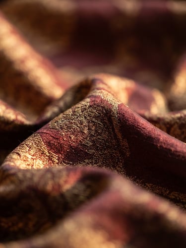 Macro photography of deep maroon and gold silk fabric draped elegantly under warm directional light, showing rich texture and weave.