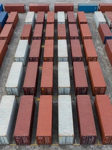 Aerial overhead shot of a freight yard — rows of shipping containers arranged in a grid under flat overcast daylight, container numbers visible on top surfaces, no personnel, operational scale evident