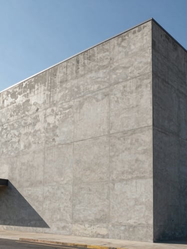 Wide-angle architectural elevation of a massive concrete commercial building under harsh midday sun, casting deep geometric shadows across its raw-cement gray facade.