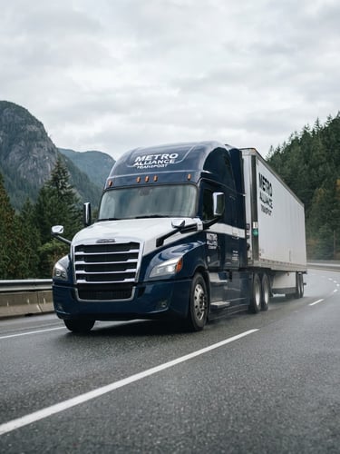 Low-angle heroic shot of a branded Metro Alliance Transport Freightliner Cascadia Class 8 truck on a scenic British Columbia highway, overcast Pacific Northwest sky, crisp morning daylight, 35mm commercial photography