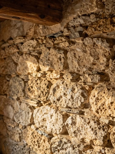 Detailed close-up of a rustic stone wall made of Caen limestone, warm sunlight grazing the texture, exposed oak beam in a cozy Normandy farmhouse interior, soft focus, editorial photography
