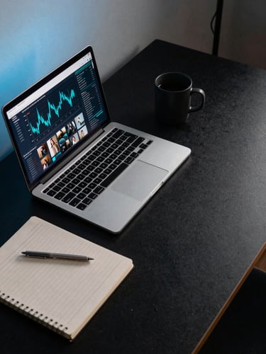 Wide overhead shot of a minimal desk workspace — open laptop showing a social media analytics dashboard with cyan data lines, a ruled notebook, and a matte black coffee mug, lit by cool blue-hour studio strobe from the left, dark surface below, generous negative space to the right