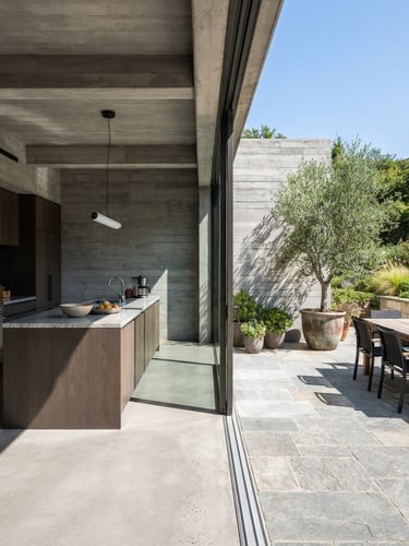 Sharp architectural photography of a modern home with a clean transition from a concrete-framed kitchen to a stone-paved garden patio, bright natural daylight, high-contrast shadows.
