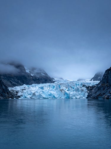 Wide cinematic view of a quiet Alaskan fjord at blue hour, deep blue waters reflecting massive glaciers, mist hanging low, expansive dark sky with negative space on the left.