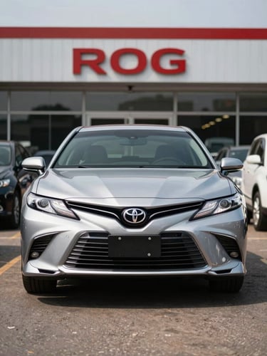 Wide three-quarter front view of a clean silver Toyota Camry on a bright daylight dealership forecourt in Accra, tarmac visible, showroom signage softly out of focus in background, no people