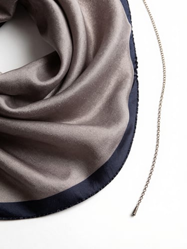 Close overhead shot of a silk scarf partially unfolded on a white surface, the weave texture and hand-rolled hem sharp under controlled studio light, a single strand of fine chain resting beside it showing scale, no hands visible, magazine-quality stillness