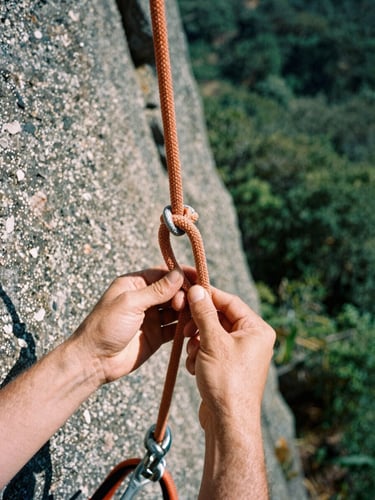 Tight macro shot of hands tying a figure-eight knot on a dynamic safety orange climbing rope, textured grey rock wall in the background, harsh natural sunlight, rugged outdoor photography, 35mm film.