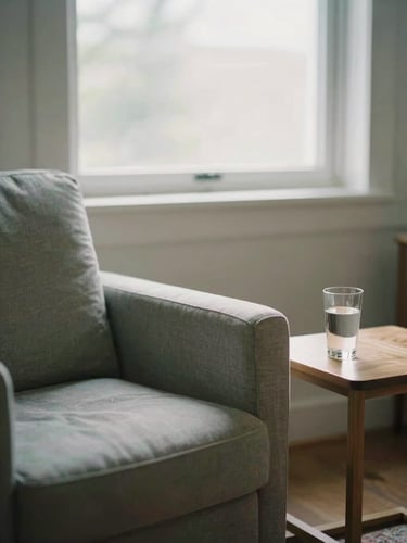 A quiet, sunlit therapist's consulting room with a comfortable fabric armchair, a small wooden side table with a glass of water, soft diffused overcast daylight coming through a window, shallow depth of field, 35mm.
