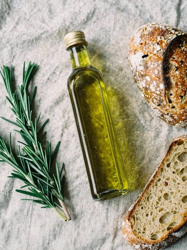 Overhead table spread of Greek olive oil, raw rosemary, and sourdough bread on a rustic linen cloth, soft natural side-light, rich crimson tones, 35mm film