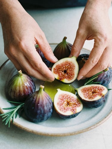 A close-up shot of hands arranging fresh figs, rosemary, and Greek olive oil on a rustic ceramic plate, soft natural daylight, moody shadows, 35mm film.