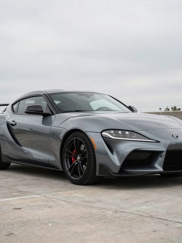 A clean, dark metallic grey sports car parked at a 3/4 angle in soft overcast Texas daylight, neutral concrete background, high-contrast reflections on the body panels.