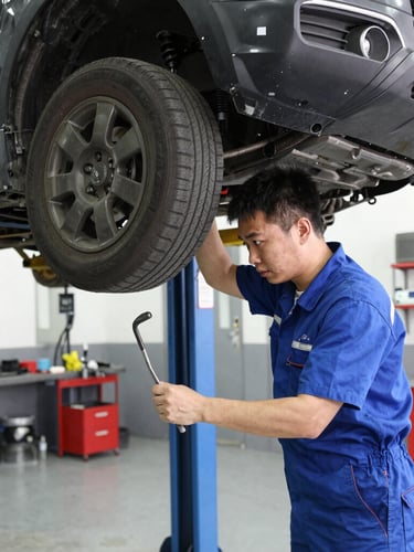 Straight-on eye-level view of a clean, modern auto workshop bay with LED lighting, a car raised on a lift, and a mechanic focused on inspecting the braking system.