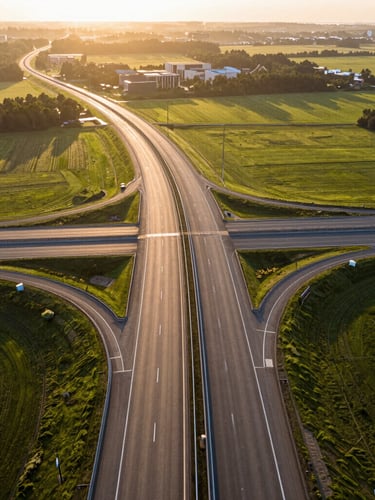 Wide aerial view of a modern highway intersection at golden hour, clean asphalt roads cutting through planned green fields, warm sunlight glinting off the lanes