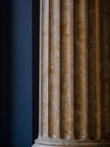 Close-up of a classical architectural column, made of warm-toned marble, with soft, directional window light highlighting its texture, deep navy and warm brass tones, shallow depth of field, 50mm