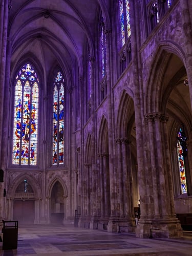 Wide low-angle view of gothic cathedral nave interior, deep violet ambient light filtering through stained glass windows far left, stone vaulted arches receding into shadow on the right, mist pooling along the floor, solemnly cinematic