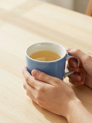 Eye-level 35mm photograph of hands holding a warm ceramic mug on a light wooden table, soft morning sunlight casting gentle shadows.