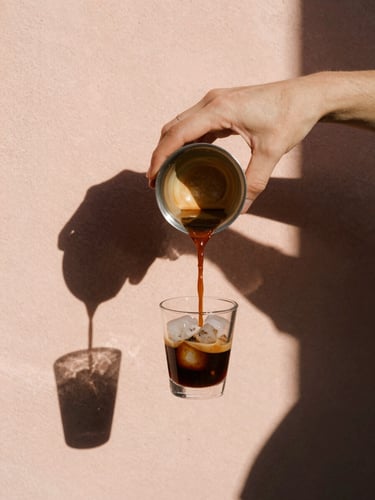 Close-up of hands pouring dark espresso over ice, bright sunlight, sharp shadow lines on peach-colored plaster, 35mm film