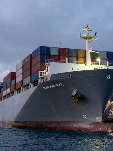 Low-angle heroic shot of a massive container ship docked at dusk, harsh industrial lighting, steel gray tones, cold blue ocean water in the foreground, high-contrast overcast sky.
