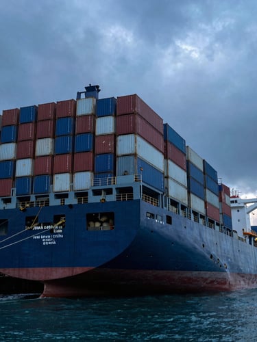 Low-angle heroic shot of a massive steel container ship docked at an industrial port during dramatic dawn, high-contrast overcast light, cold blue ocean tones.