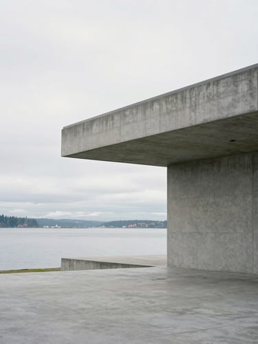 A wide crop of minimalist studio architecture on Bainbridge Island, clean concrete lines meeting cool grey water under a bright overcast sky, generous negative space.
