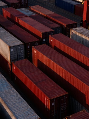 Stark aerial view of a massive container shipping hub at dusk, sharp directional light casting long shadows on deep crimson containers, 35mm film grain