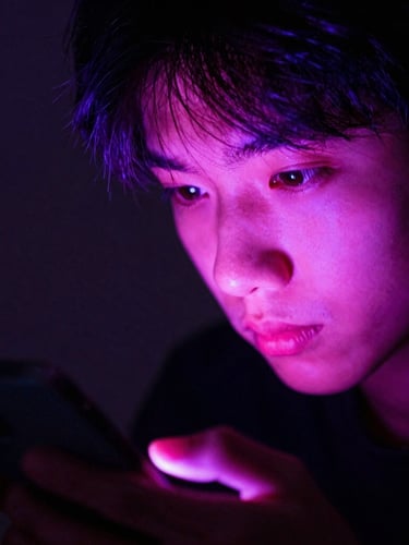 Close-up of a young creator's face illuminated by the bright pink and violet glow of a smartphone screen, dark background, intense focused expression, high contrast.