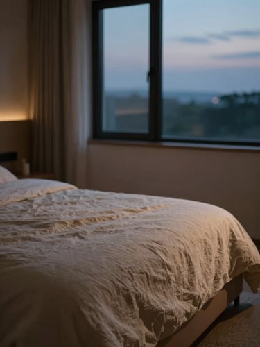 A quiet, minimalist bedroom sanctuary at twilight, soft amber evening light filtering through a window onto a perfectly made bed with organic linen textures, shallow depth of field, 35mm.