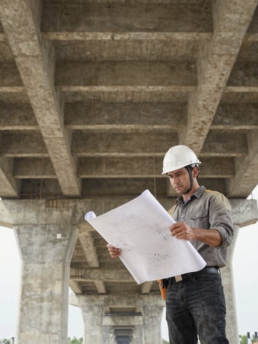 Low angle heroic shot of a civil engineer in a white hardhat reviewing blueprints on a massive concrete bridge construction site under high-contrast daylight