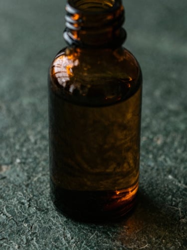 Macro overhead shot of dark amber glass apothecary bottle on a textured charcoal stone surface, rich copper-gold light catching the glass, deep forest green shadows, moody 35mm photography