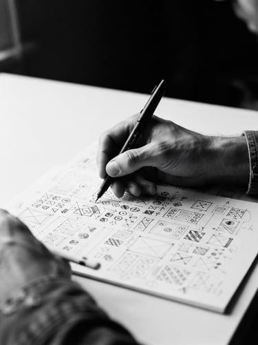 Close-up of hands sketching a visual identity system on a clean desk, natural window side-lighting, high contrast black and white, editorial portrait photography style
