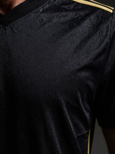 Close-up macro shot of an official football jersey fabric, showing intricate texture and stitching, with a subtle gold thread detail, moody editorial sports photography, dramatic high-contrast studio lighting, deep black and gold tones, sharp details, 35mm