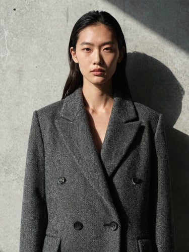A stark high-contrast portrait of a model in a structured wool coat, casting a sharp geometric shadow on a raw concrete wall, natural overcast light.