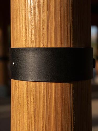 Extreme close-up of a hand-hewn cedar column base meeting a matte black iron bracket, golden side-light raking across the grain to reveal deep wood texture, tight architectural framing, no people, warm amber tones against deep shadow