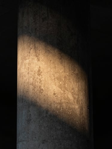 Dramatic vertical portrait of a monolithic concrete column, a sharp diagonal beam of warm amber light grazing the rough texture, deep black shadows on the left, 35mm lens