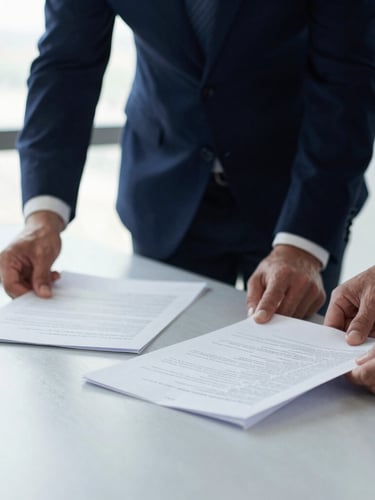 A close-up of two professionals reviewing physical documents on a light slate gray table, bright natural side light from a window, shallow depth of field, professional and structured atmosphere.