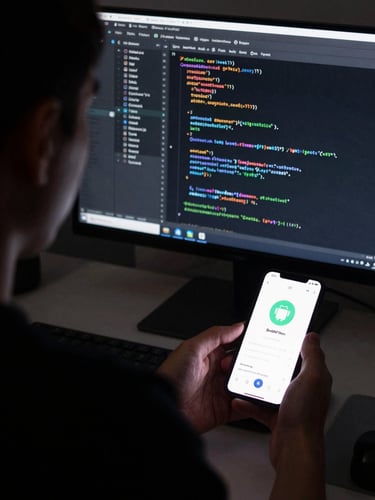 Tight over-the-shoulder perspective of a developer inspecting native Swift code on a dark monitor next to a physical Android device displaying a live app layout