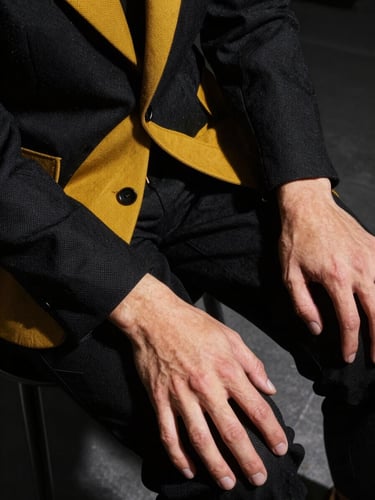 Extreme close-up overhead shot of a styled artist's torso and hands against a dark studio floor, structured jacket with visible texture detail under a focused strobe spotlight, hands resting on knees, sharp angular shadows, high contrast black and mustard-yellow fabric accents
