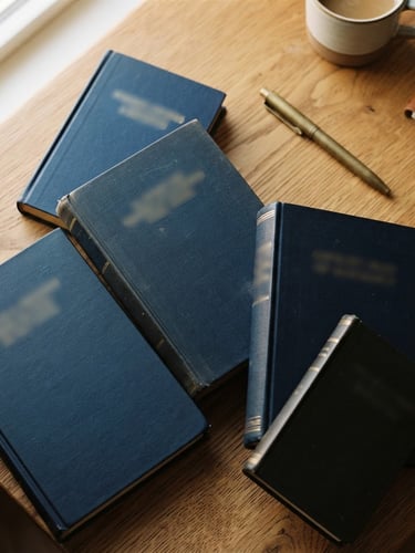 Overhead flatlay of thick academic textbooks and a brass pen on a clean wooden desk, warm daylight, shallow depth of field