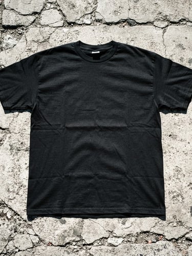 Raw flash photography of a heavy black cotton t-shirt flat-lay on cracked concrete, harsh direct sunlight, high-contrast shadows.