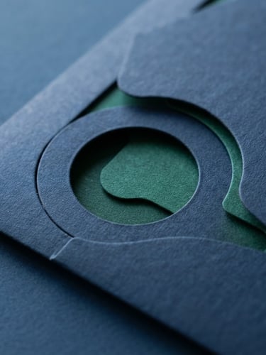 Macro close-up of a high-quality physical direct mail piece with sharp custom die-cuts, textured premium paper, resting on a clean dark blue desk under bright natural daylight.