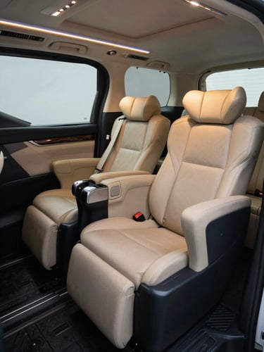 Interior of a Toyota Alphard executive rear cabin, ottoman footrest extended, cream and charcoal fabric seats with precise stitching, ambient cabin lighting glowing softly along the ceiling rail, cool-neutral light filtering through tinted rear window, no passengers, composed and still
