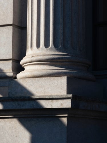 Macro shot of a classic stone pillar of an institutional financial building, sharp geometric shadows, soft morning window light, deep navy tones, editorial 35mm photography.