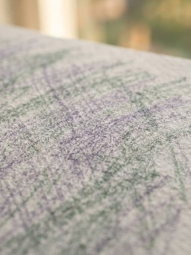 Macro close-up of textured artist paper with soft colored-pencil strokes in lavender and sage, sunlit studio background, extremely shallow depth of field