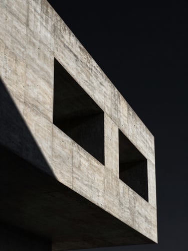 Dramatic vertical crop of hard-edged raw concrete architecture against a pitch-black sky, high-contrast studio key light casting deep geometric shadows.