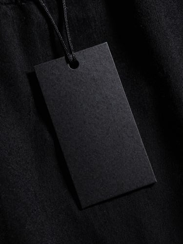 High-fashion vertical crop shot of a stark black garment tag, hard studio key light, deep unforgiving shadows.