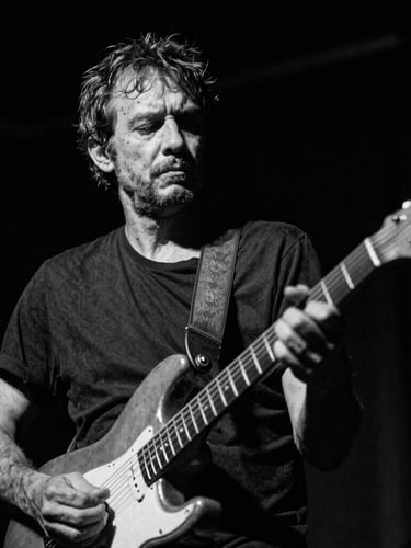 Candid black and white portrait of a weathered male musician with a guitar, low-key dramatic stage lighting, deep shadows, 35mm film grain