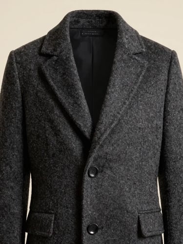 Polished editorial model wearing a structured charcoal wool coat, high-contrast studio lighting, warm cream background, sharp focus, 35mm lens