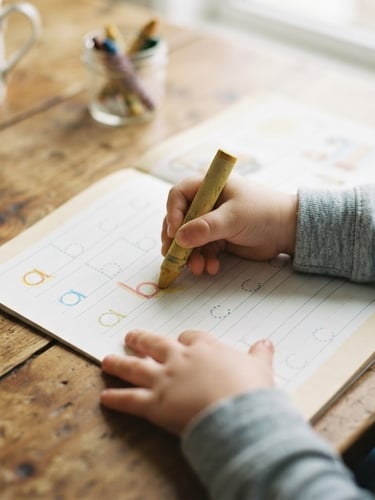 Overhead shot of children's hands holding a thick crayon, tracing letters on a printed workbook page, on a warm wooden kitchen table, soft diffused morning daylight, gentle muted pastel tones, shallow depth of field, 35mm film look