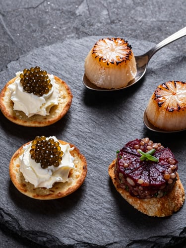 Close-up overhead of four passed appetizers arranged on a dark lacquered tray — blini topped with crème fraîche and caviar, a seared scallop on spoon, a tartare bite on crisp, a roasted beet crostini — amber studio light from above, deep shadow between pieces, slate surface